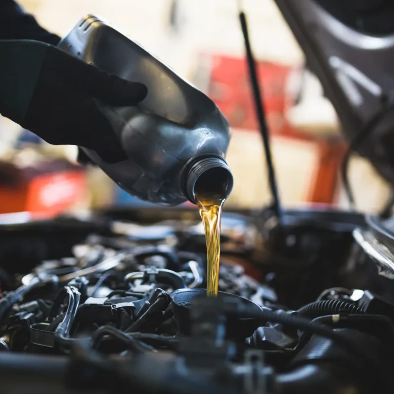 A mechanic wearing black gloves pours golden motor oil from a bottle into the oil filler opening of a car engine.