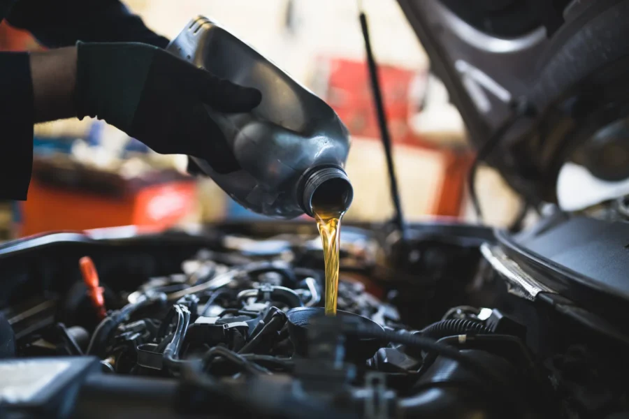 A mechanic wearing black gloves pours golden motor oil from a bottle into the oil filler opening of a car engine.