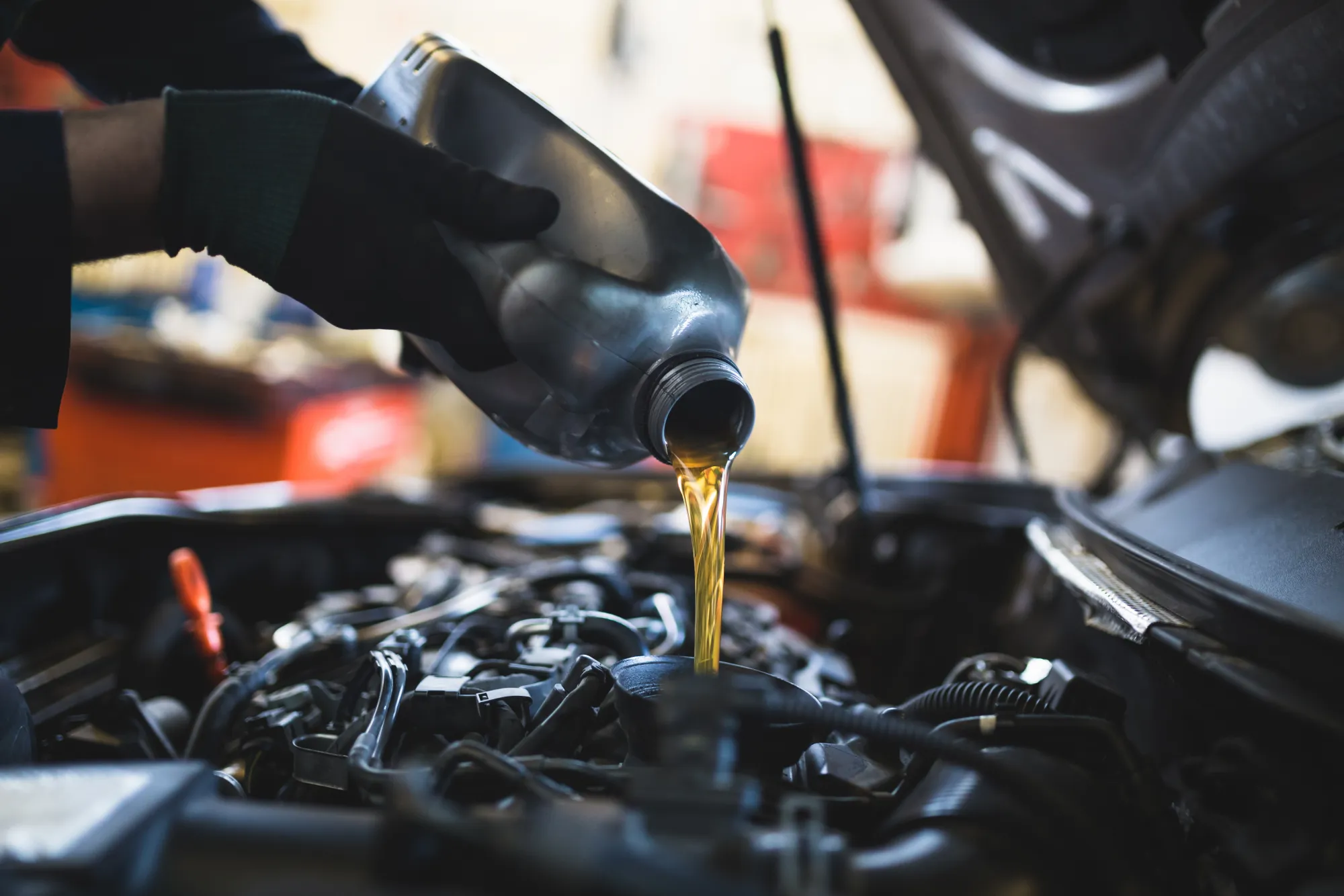 A mechanic wearing black gloves pours golden motor oil from a bottle into the oil filler opening of a car engine.