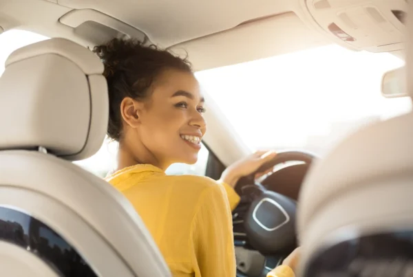 A smiling young woman in a yellow top sits behind the steering wheel of a car with sunlight coming through the windshield, looking back over her shoulder.