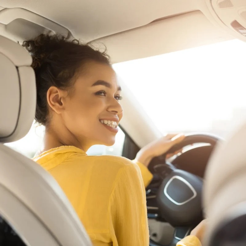 A smiling young woman in a yellow top sits behind the steering wheel of a car with sunlight coming through the windshield, looking back over her shoulder.