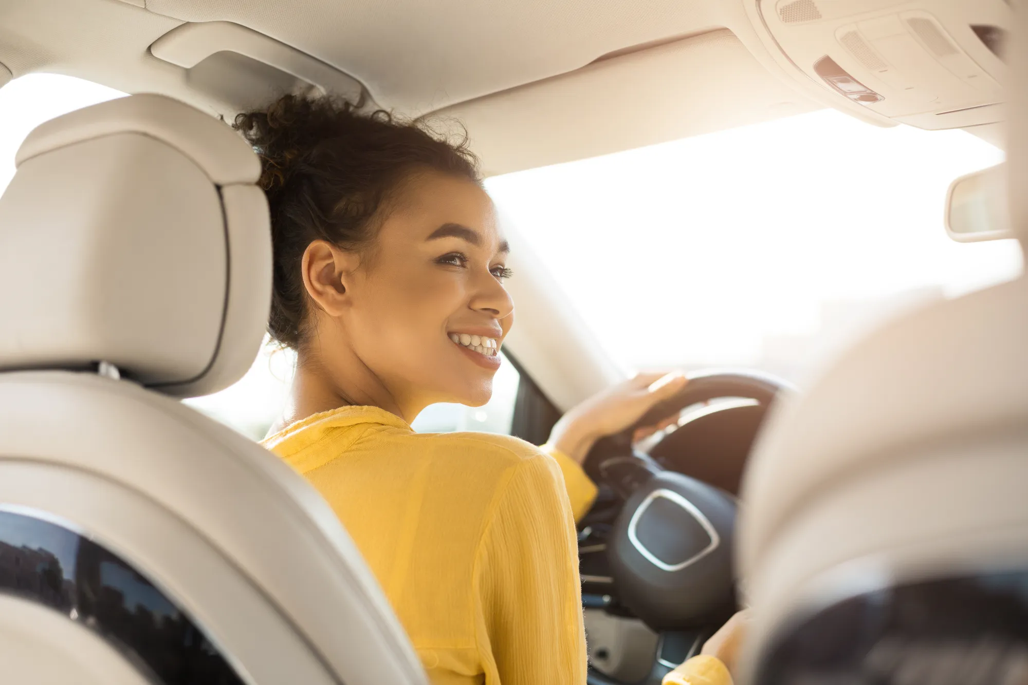 A smiling young woman in a yellow top sits behind the steering wheel of a car with sunlight coming through the windshield, looking back over her shoulder.