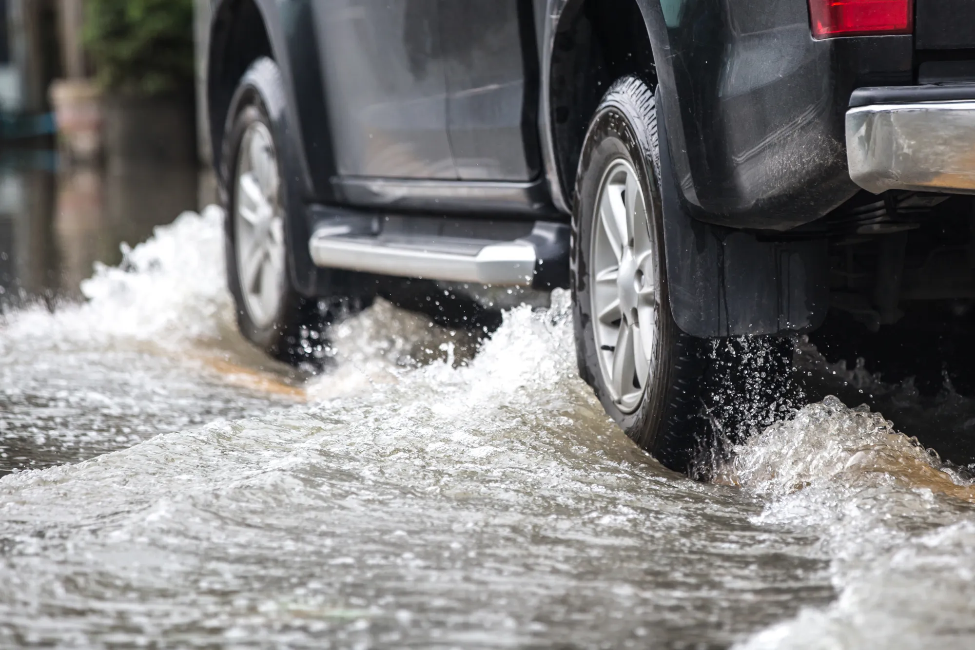 Rear tire and wheel of a dark SUV splashing through deep water on a flooded road.