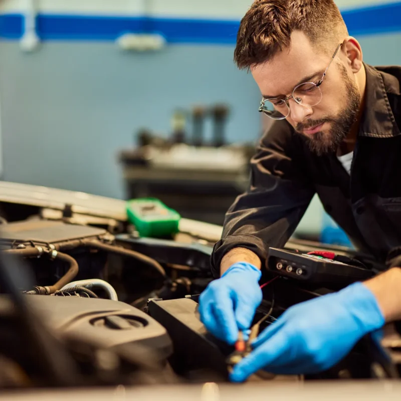 A mechanic wearing blue gloves tests a car battery with a multimeter under an open hood in an auto repair shop.