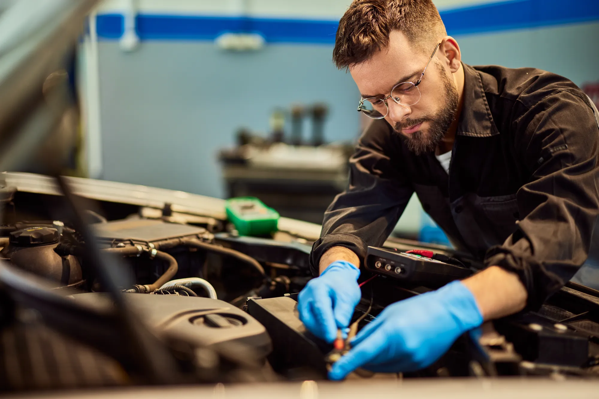A mechanic wearing blue gloves tests a car battery with a multimeter under an open hood in an auto repair shop.