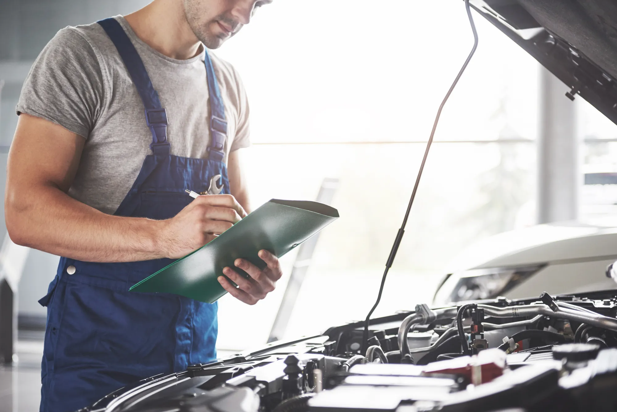 A mechanic in blue overalls writes on a clipboard while standing beside a car with its hood open in a repair shop.