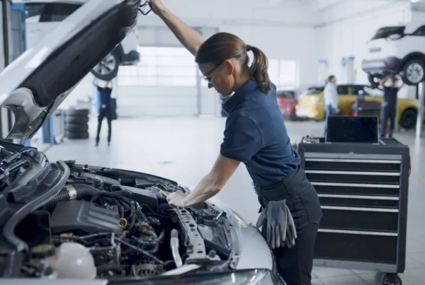 A female auto technician holds open a car's hood and reaches into the engine bay inside a bright repair shop with vehicles on lifts behind her.