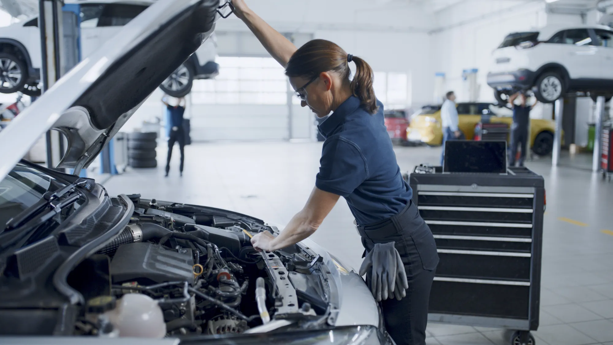 A female auto technician holds open a car's hood and reaches into the engine bay inside a bright repair shop with vehicles on lifts behind her.